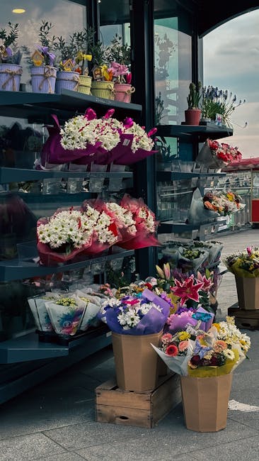 A display of fresh flower arrangements on black metal shelves, featuring bouquets of white, pink, purple, and yellow blooms wrapped in colorful paper and cellophane. The bouquets include roses, lilies