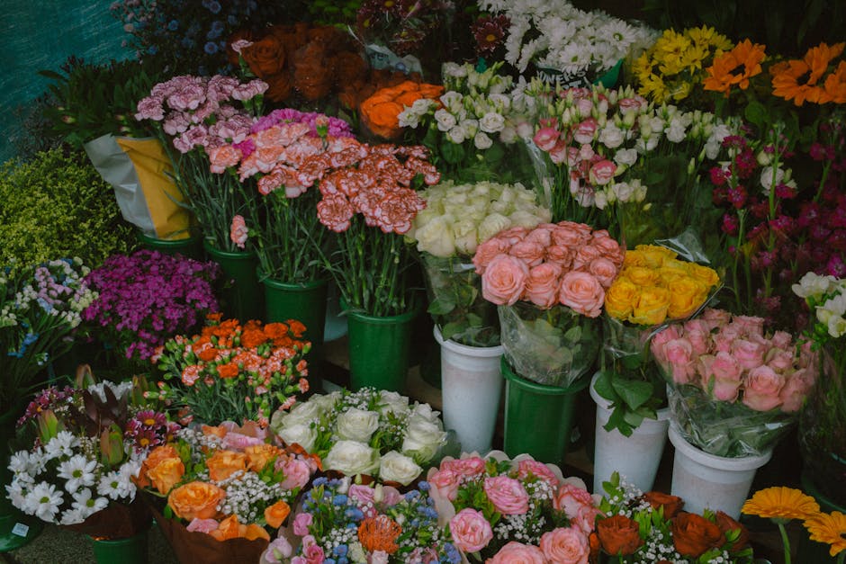 A vibrant display of fresh floral bouquets and potted plants arranged on shelves and the floor at a flower shop, including pink roses, white roses, yellow blooms, orange carnations, purple and pink ch