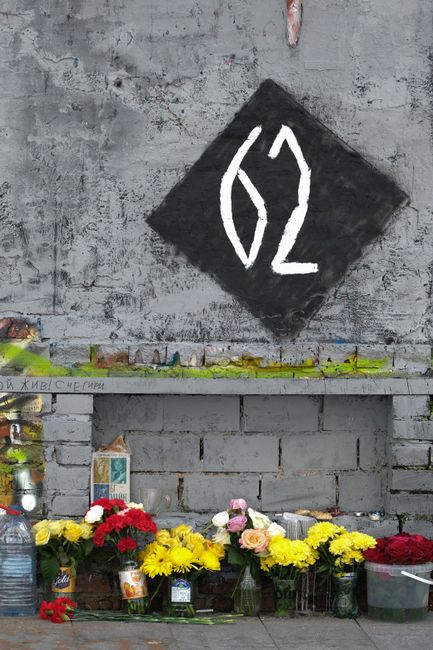 A floral tribute placed against a weathered gray brick wall beneath a black diamond-shaped sign with white stylized numbers '62'. The tribute consists of various fresh flowers, including bright yellow
