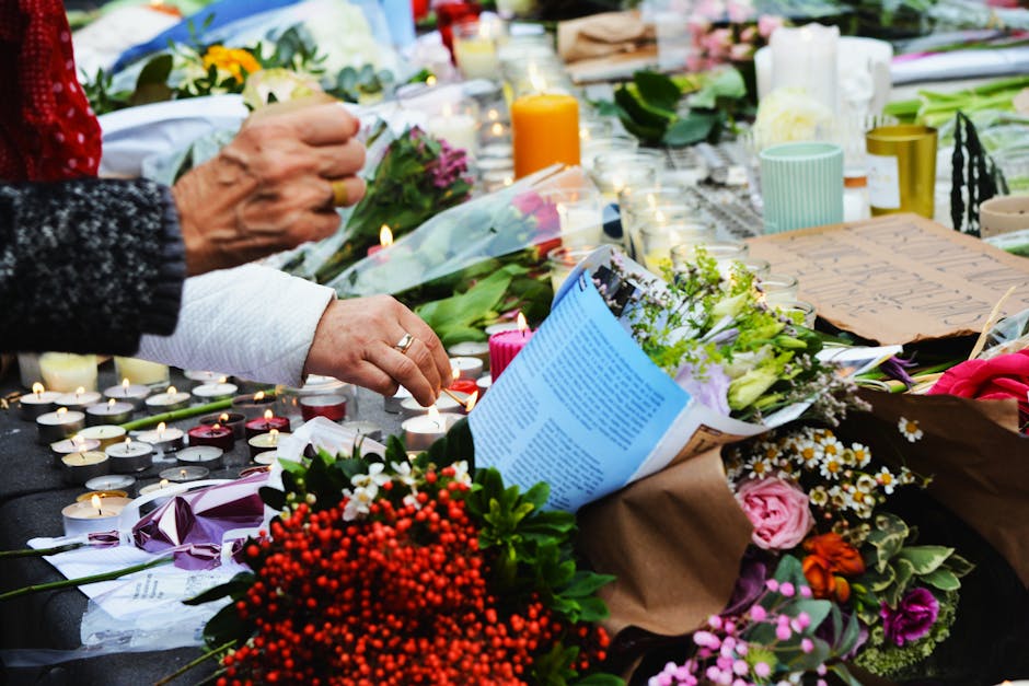 A floral tribute scene featuring a variety of fresh flowers including pink roses, white daisies, purple statice, and red berries arranged on a memorial surface. The arrangement is wrapped in brown pap