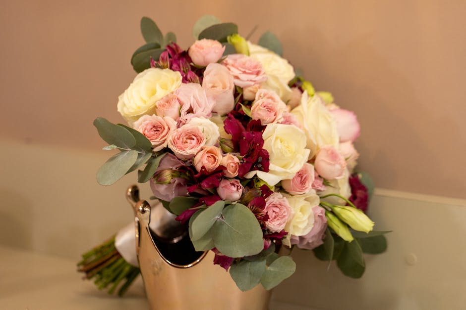 A close-up image of a wedding bouquet featuring a mix of fresh flowers, including soft pink roses, creamy white eustoma, and deep burgundy blossoms, accented with green eucalyptus leaves. The bouquet 