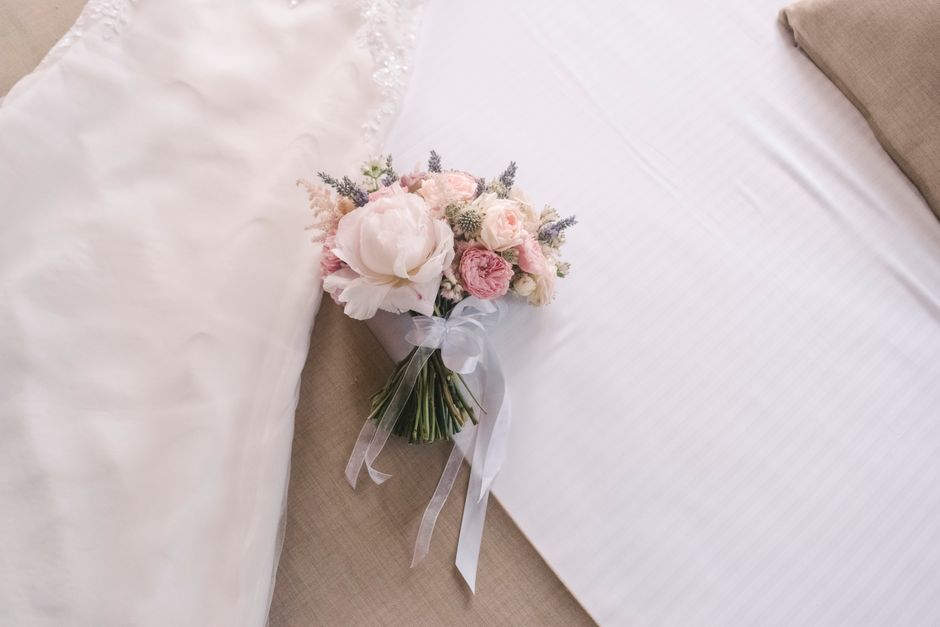 A delicate bridal bouquet featuring soft pink and white peonies, roses, and small lavender sprigs, arranged in a rounded style. The flowers are fresh with slightly textured petals and green stems, wra