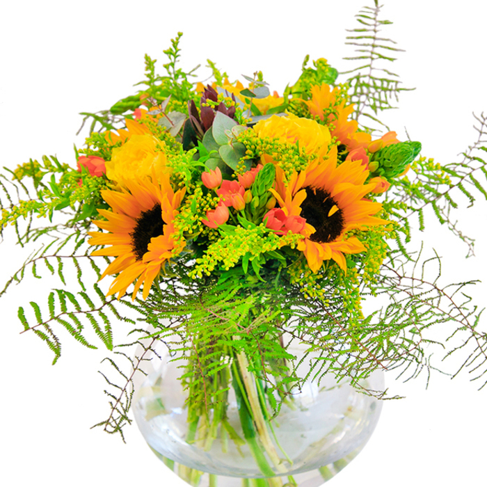 From the moment you glimpse this Beckton bouquet, it radiates exuberance and celebration with its bold pairing of sunflowers and canary-yellow roses-nature's own cheerleaders. Each sunflower head is a study in contrasts, with plush, velvety petals circling deep, chocolate-brown centres, bringing an aura of sun-soaked fields right into a Beckton home. The roses, gathered in elegant swirls of layered petals, add refinement and a tactile softness-each bloom reminiscent of the floral abundance seen in gardens along Stansfeld Road. Among the blossoms, clusters of red Hypericum berries sparkle brightly, their slick, lively skins offsetting the matte textures of their neighbours. Ferns trail gracefully over the edges, ebbing and flowing like the grasses of Beckton Corridor, while light airy sprigs of solidago scatter minuscule yellow blooms throughout, enhancing depth and offering a gentle wildness. Wisps of eucalyptus bring cool, subtle greens and a fragrant lift. Through the crystal round vase, vibrant stems intertwine in neat precision, their natural architecture on full display, roots in place and alive in clear water. Textural contrasts abound-from the suede-like sunflower to the snap of crisp greens-making this bouquet a sensory delight. The air is suffused with the scent of fresh-cut roses and a hint of open parkland, evoking summer memories and festive gatherings. This arrangement is the perfect way to mark a joyful milestone or share a little brightness across Beckton, crafted with dedication and an intimate understanding of local tastes.