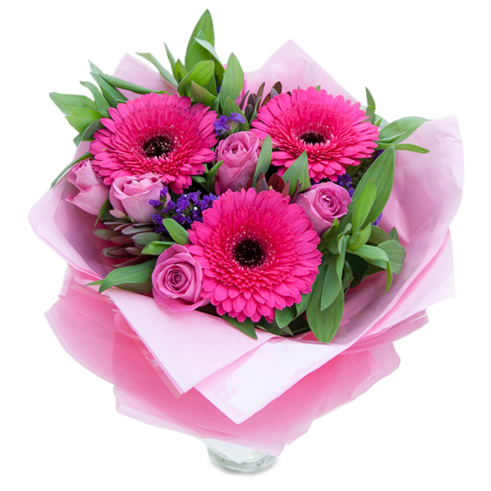 I see a bouquet that balances boldness and softness with an elegant hand: three fuchsia gerbera daisies dominate the view, their saturated petals bursting outward and framed by distinct dark centres that provide striking contrast. Around them, roses in a muted lavender-pink or dusty rose sit like soft spirals, their velvety layers conveying intimacy and care. Scattered throughout are petite clusters of purple statice or limonium, adding delicate, papery texture and a whisper of cool colour between the larger blooms. The greens-broad, glossy leaves resembling salal or eucalyptus-form a verdant backdrop that lifts the pinks and provides a sense of natural fullness and slight herbal freshness. The arrangement is enveloped in a pale pink tissue lining and finished with an outer layer of translucent fuchsia-pink cellophane that shimmers faintly and ties the palette together with chic cohesion.
The top rim of a clear glass vase is just visible at the lower edge, suggesting the bouquet has been prepared for immediate display on a dining table or mantel. In soft morning light the petals catch and reflect subtle highlights; the fragrance is understated and pleasant, roses giving a gentle perfume while gerberas bring a clean, green sweetness. Picture this arriving as a thoughtful gift after a stroll near Beckton District Park or left as a cheerful centrepiece for a local celebration. The composition reflects experienced floral technique, aiming to uplift and celebrate with freshness and refined colour harmony tailored to Beckton homes and occasions.